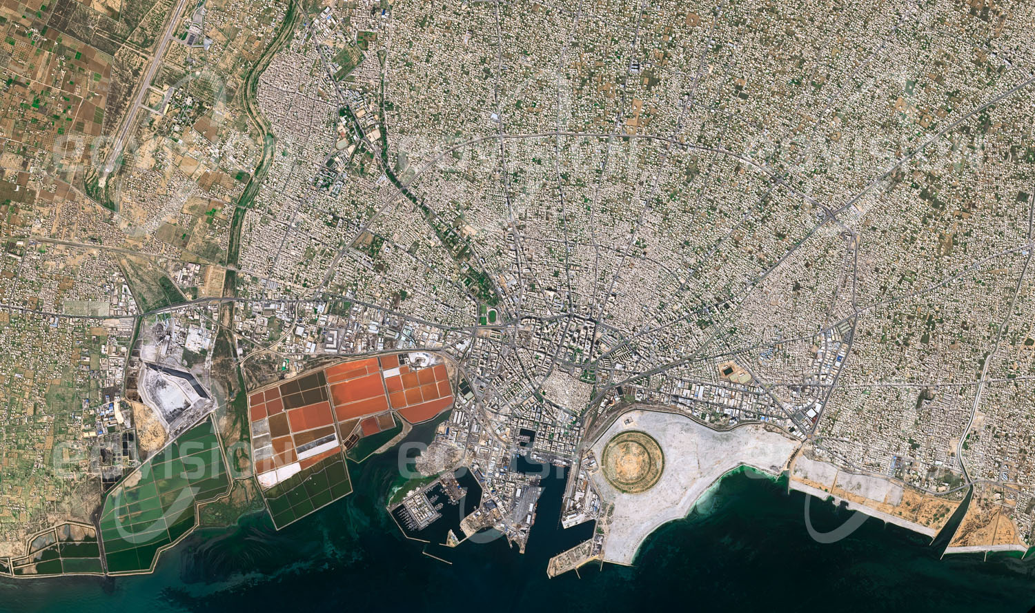 Das Satellitenbild 'SFAX, Tunesien' ist dem Bildband "Cities - Brennpunkte der Menschheit" entnommen. Sfax, mit 330.000 Einwohnern zweitgrößte Stadt Tunesiens, fällt im Satellitenbild durch seinen konzentrischen Aufbau auf. Vom alten Zentrum mit seinen kleinen Gebäuden und engen Gassen verlaufen die Hauptstraßen sternförmig nach außen, wo sie spinnennetzartig durch Ringstraßen verbunden sind, wobei nach außen die Bebauung lockerer wird. In der kreisförmigen Struktur am Meeresufer wird eine ausgedehnte Parkanlage errichtet. Sie liegt im Stadtentwicklungsgebiet Taparura, das nach Fertigstellung 50.000 Bewohner aufnehmen soll.