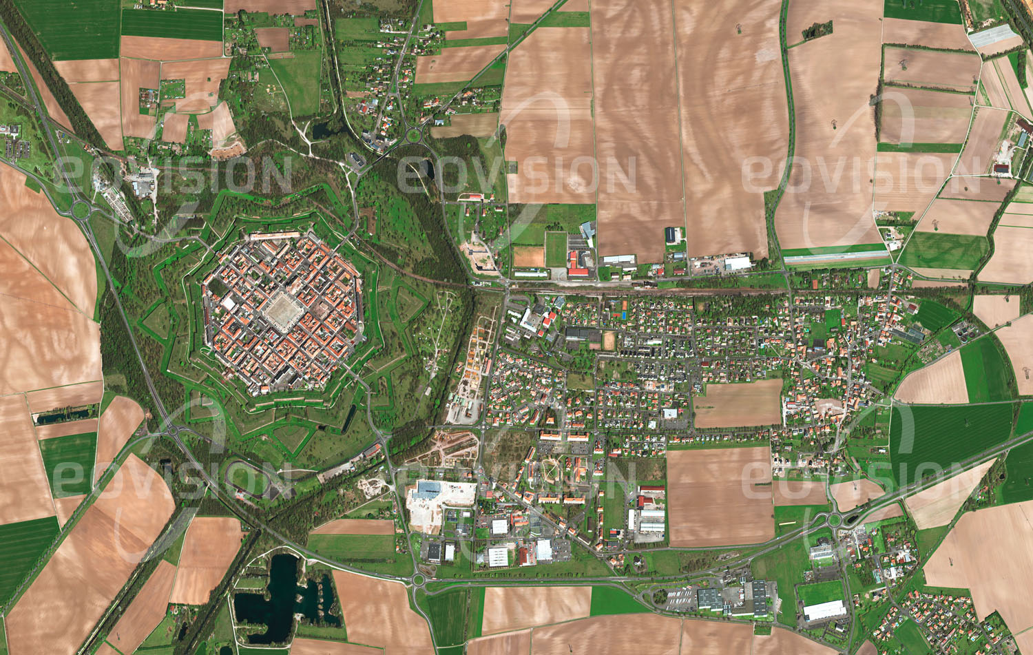 Das Satellitenbild 'NEUF-BRISACH, Frankreich' ist dem Bildband "Europa - Kontinent der Vielfalt" entnommen. Neuf-Brisach wurde in den Jahren um 1700 nahe dem Rhein errichtet, da der Fluss nach Beendigung des pfälzischen Erbfolgekriegs die neue Grenze zwischen Frankreich und dem Heiligen Römischen Reich bildete. Neuf-Brisach wurde vom französischen Festungsbauer Vauban als Ideale Stadt geplant: rechtwinkelige Gassen auf einem achteckigen Grundriss sowie die vorgelagerten Befestigungsanlagen vermitteln einen Eindruck hoher Symmetrie. Mangels beruflicher Möglichkeiten in der Kleinstadt pendeln die meisten der etwa 2000 Einwohner in die benachbarten Städte aus.n