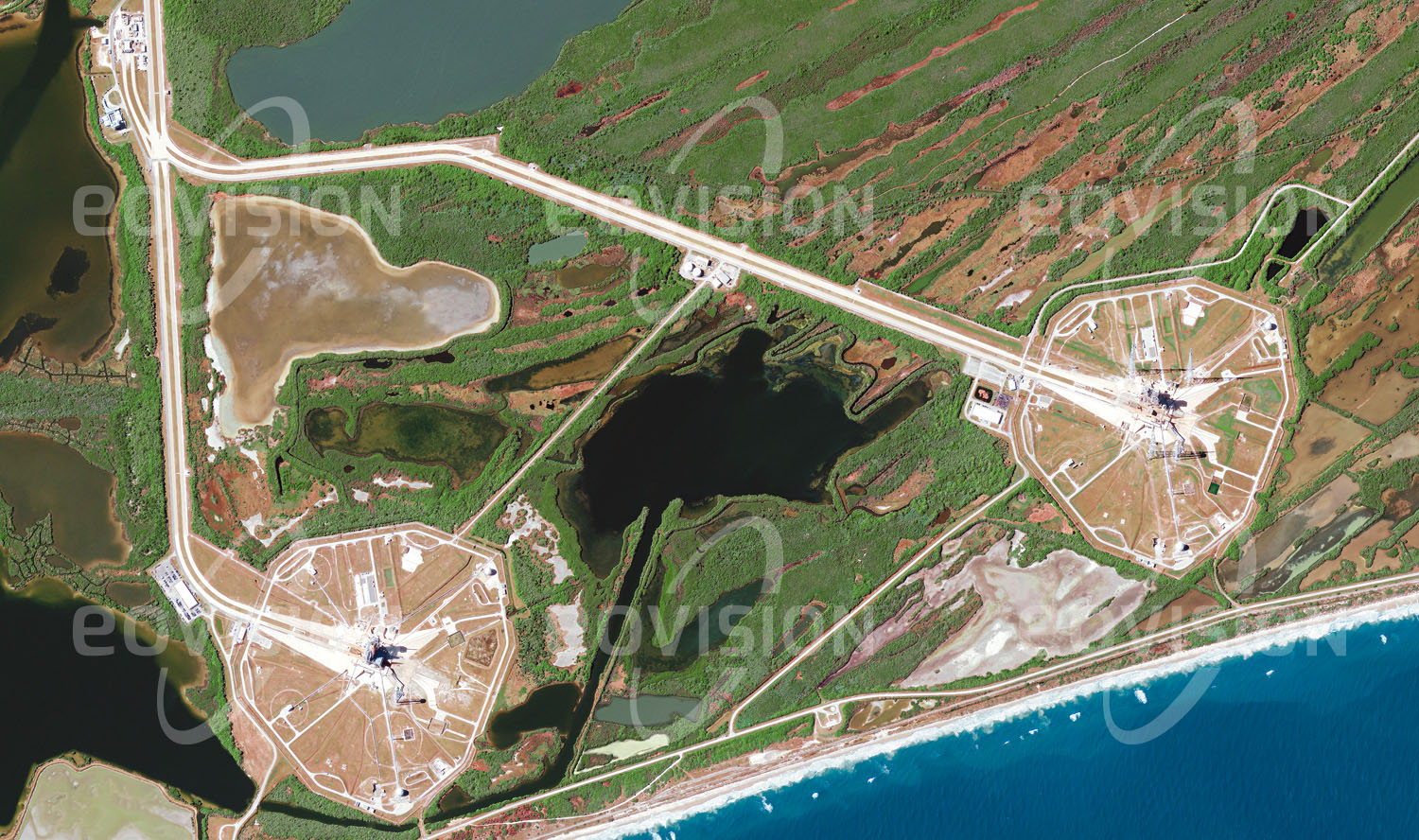 Das Satellitenbild 'CAPE CANAVERAL, USA' ist dem Bildband "Human Footprint - Satellitenbilder dokumentieren menschliches Handeln" entnommen. Direkt an der Atlantikküste liegen die beiden Raketenstartrampen mit den Starttürmen, die jeweils von vier Blitzableitern sowie je zwei Tanks für Sauerstoff und Wasserstoff umgeben sind.