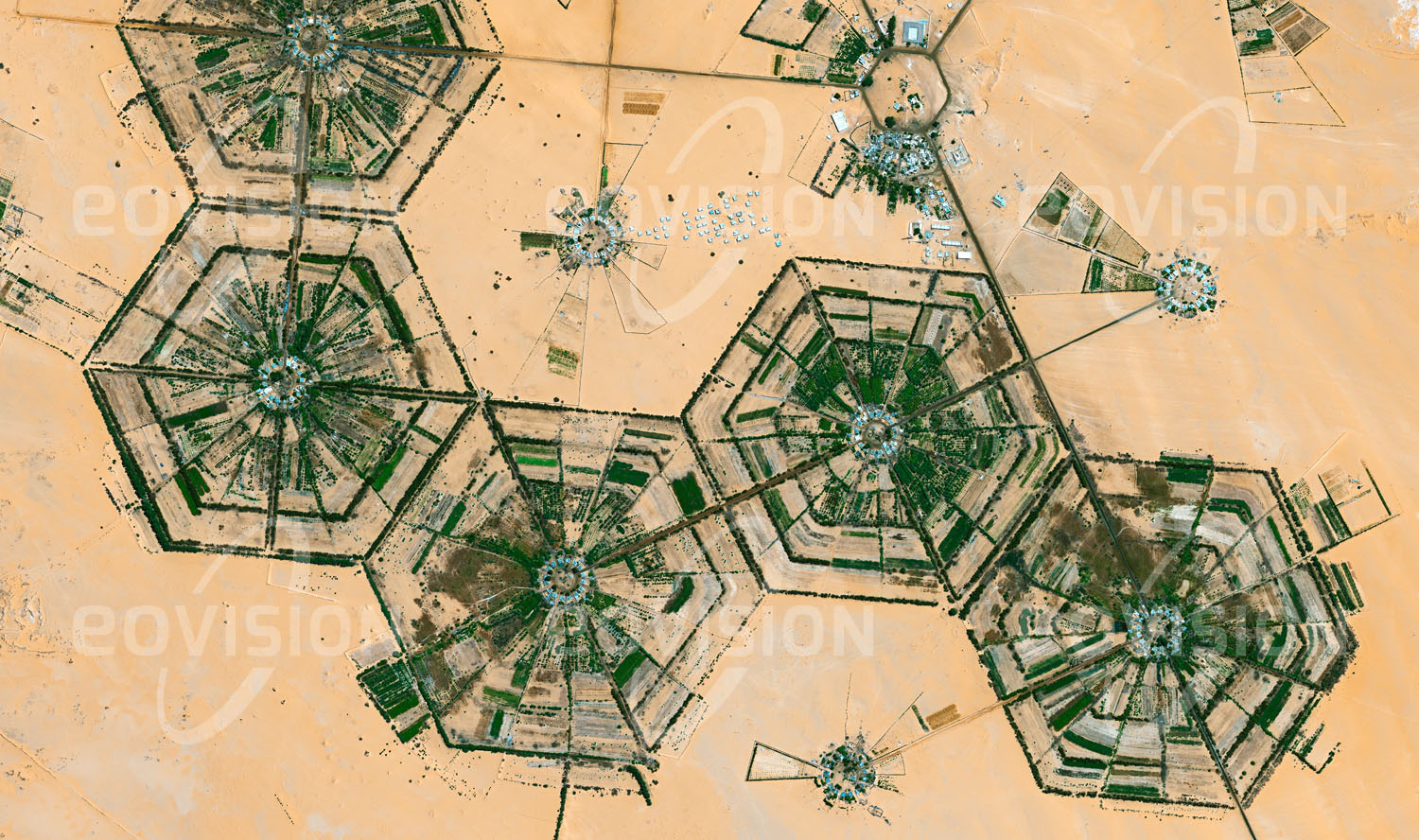 Das Satellitenbild 'AL KUFRAH, Libyen' ist dem Bildband "Human Footprint - Satellitenbilder dokumentieren menschliches Handeln" entnommen. Das fossile Grundwasser, mit dem die Felder um Al Kufrah bewässert werden, wird wegen des trockenen Klimas kaum nachgebildet, sodass ein Ende der Wasservorräte absehbar ist. Nahe den Mittelpunkten der abgebildeten sechseckigen Strukturen liegen Wohn- und Wirtschaftsgebäude, deren Anbauflächen für Obst und Gemüse innerhalb der Sektoren nach außen angeordnet sind. Die bewirtschafteten Flächen werden an der Außengrenze durch Busch- und Baumreihen vor dem Wüsten-sand geschützt.