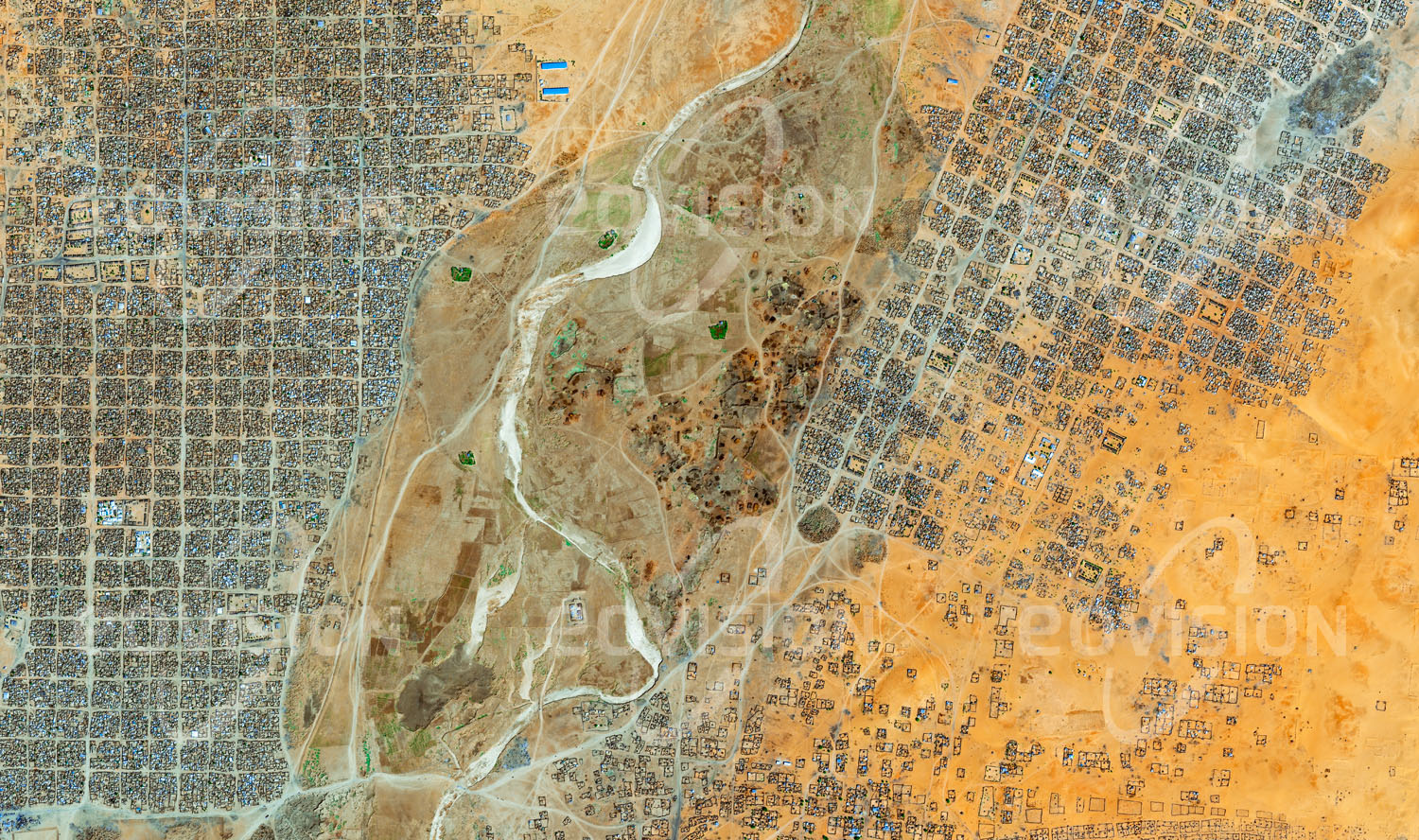 Das Satellitenbild 'AL FĀSHIR, Sudan' ist dem Bildband "Human Footprint - Satellitenbilder dokumentieren menschliches Handeln" entnommen. Als Folge des Darfur-Konflikts, der ab 2003 den Sudan erschütterte und annähernd 300.000 Todesopfer forderte, verloren etwa fünf Millionen Menschen ihre Heimat und fanden in Flüchtlingslagern Zuflucht. So entstand auch im Norden der Stadt al-Fashir im Winter 2004/05 der erste Teil des Flücht­lingslagers Abu Shouk, eine Erweiterung nach Osten folgte in den Jahren danach. Der regelmäßige Aufbau des innerhalb weniger Jahre entstandenen Lagers unterscheidet sich deutlich von der gewachsenen Struktur der ursprünglichen Siedlung.