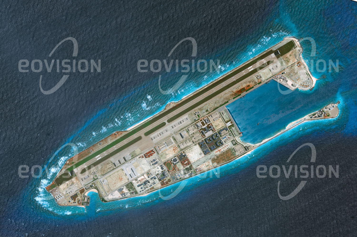 Das Satellitenbild 'FIERY CROSS REEF 31May2014,' ist dem Bildband "New Human Footprint - Unsere Welt im Umbruch" entnommen. Die beiden Satellitenbilder vom 31. Mai 2014 (oben) und vom 14. Juni 2018 (unten) verdeutlichen, welchen Aufwand Staaten zu treiben gewillt sind, um ihre Interessen zu wahren. Innerhalb weniger Jahre wurde bei den Spratly-Inseln im Südchinesischen Meer ein Korallenriff in eine Insel umgewandelt, auf der eine kleine Stadt samt Sportanlagen mit Fußballplatz und Tennisplätzen errichtet wurde. Sie beherbergt eine Militärgarnison, die über einen eigenen Flughafen mit einem 3,3 km langen Landestreifen verfügt.