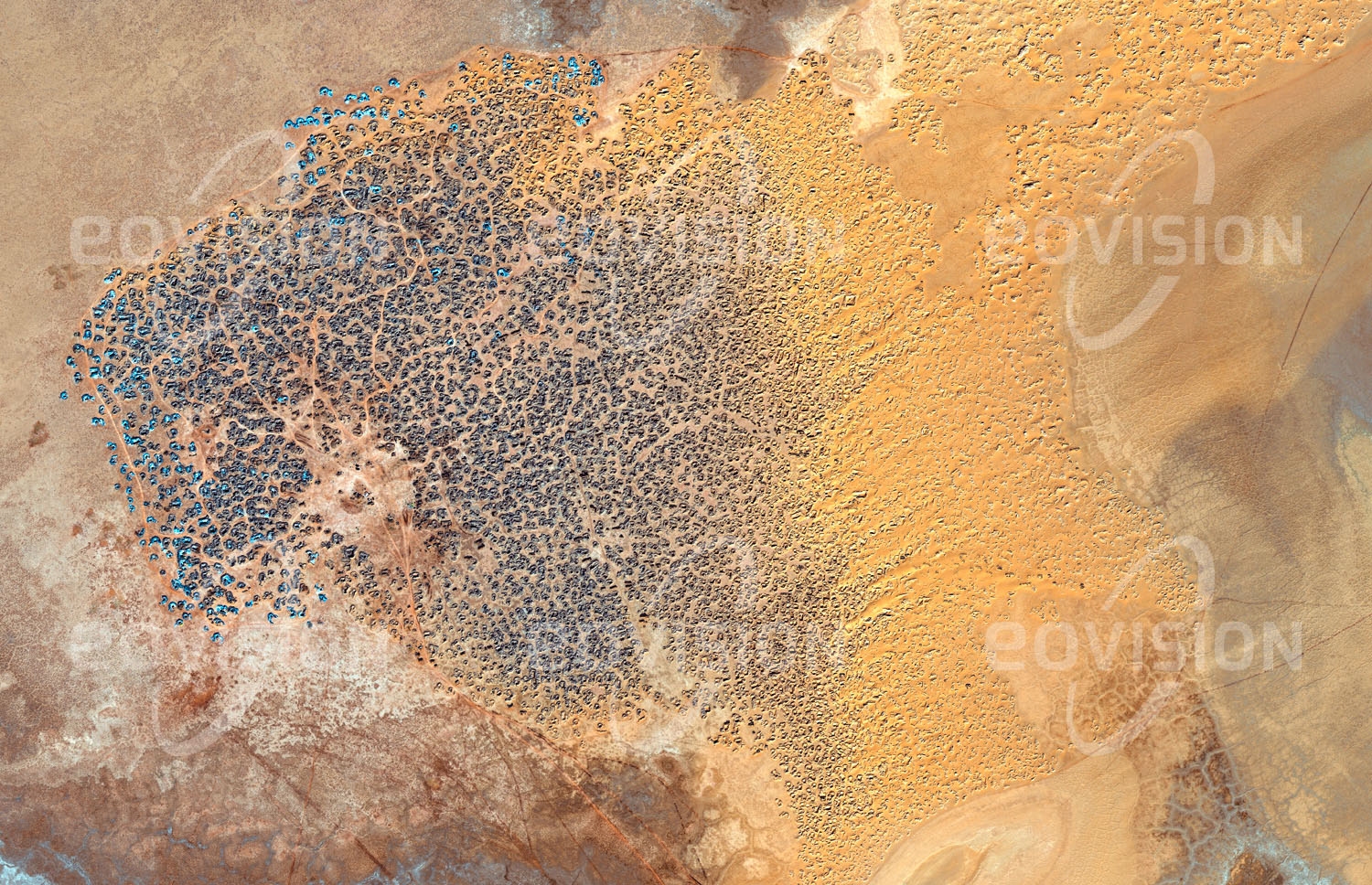 Das Satellitenbild 'TAOUDENNI, Mali' ist dem Bildband "Wüsten - Lebensraum der Extreme" entnommen. Etwa ab dem Ende des 16. Jahrhunderts wurde nahe der Stadt Taoudenni Salz gewonnen. Heute hat sich die Salzgewinnung etwa 10 Kilometer weiter in den Südwesten verschoben. Hier liegt einige Meter unter der Oberfläche eine hochwertige Salzschicht. Mit einfachsten Mitteln werden hunderte bis zu vier Meter tiefe Löcher in den Sand und Lehm gegraben. Aus der darunter liegenden Salzschicht werden Platten geschlagen, die zu 30 Kilogramm schweren Salzbarren bearbeitet werden. Karawanen transportieren diese nach Timbuktu.